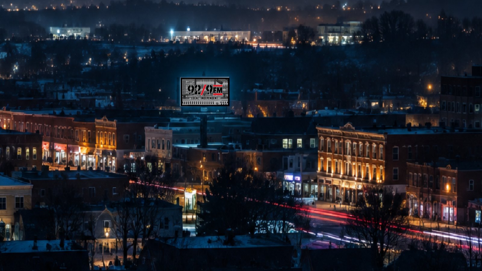 A small city at night seen from a hillside: rows of lit brick storefronts and a church steeple below a dark ridge of distant lights, red and white traffic trails streaking along the main street, and a lit billboard on a rooftop above the block reading 92.9 FM, local, independent.