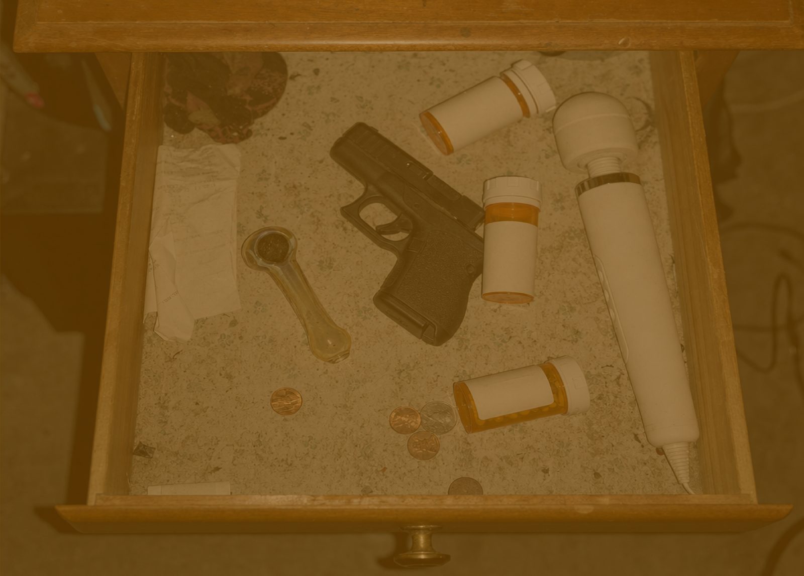 An open wooden nightstand drawer photographed from above, its contents laid bare on the liner: unlabeled prescription bottles, a handgun, a glass pipe, a personal massager, a folded paper and loose change.