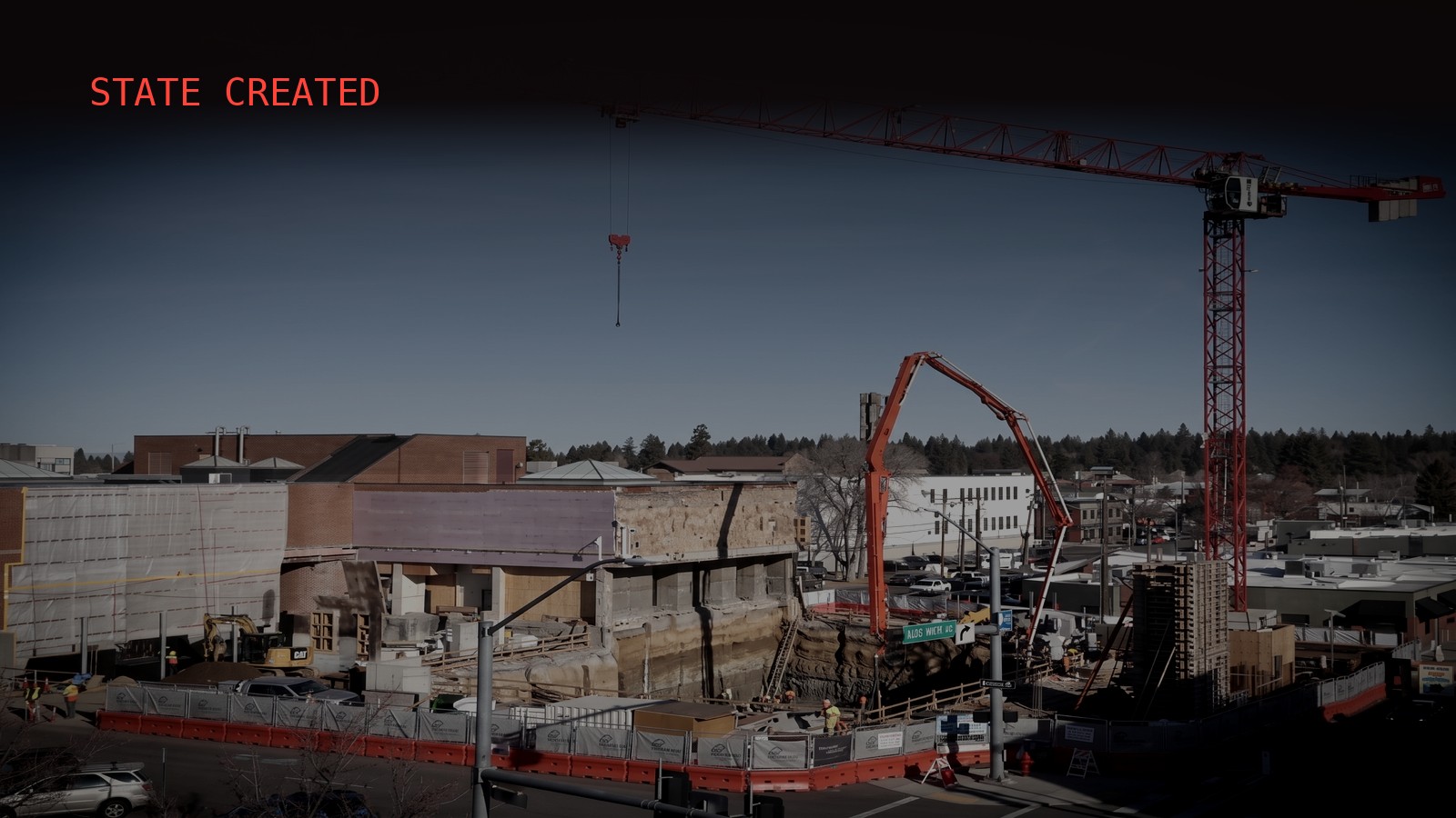 A city block under construction at dusk: a red tower crane stands over a deep open excavation pit ringed by orange safety fencing, a concrete pump arm reaching down into it, and workers in high-visibility vests spread along the edge.
