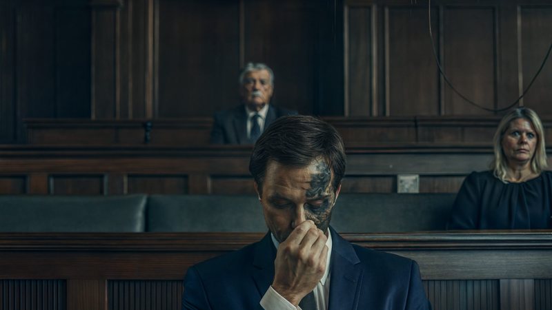 A man in a navy suit seated alone at a courtroom table, head bowed with a hand to his face, exposed circuitry showing beneath the skin on one side of his face while a judge and another figure look on.