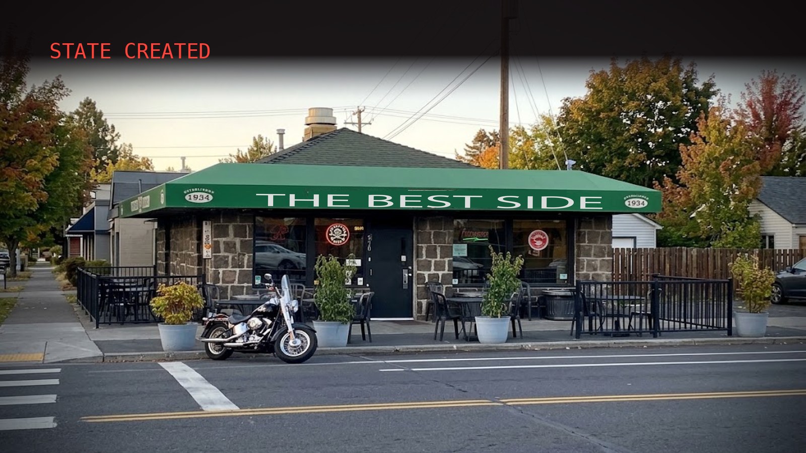 A small stone-walled corner tavern under a green awning reading &ldquo;The Best Side,&rdquo; a black motorcycle parked at the curb out front, autumn trees and an evening sky behind it.